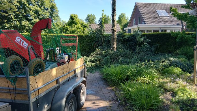 Een aanhangwagen met tuingereedschap staat op een pad, omringd door groen.